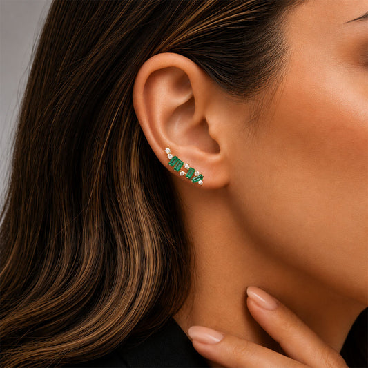 A close-up of a woman's ear wearing a curved ear cuff earring with baguette colombian emeralds and small diamonds in a 18k yellow gold setting.