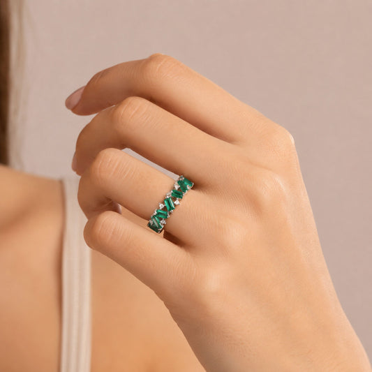 A close-up of a hand wearing a white gold band ring set with baguette colombian emeralds and small diamonds, against a soft beige background.