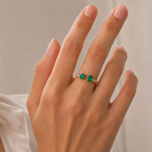 A close-up of a hand wearing a f round and emerald-cut Colombian emeralds on open toi et moi ring, Duet Ring by GOHI Soiree Collection. A 18k fairmiend yellow gold ring featuring a large rectangular emerald-cut Colombian emerald gemstone, a smaller round emerald-cut colombian emerald on the opposite side, and a row of round brilliant-cut diamonds along the band.