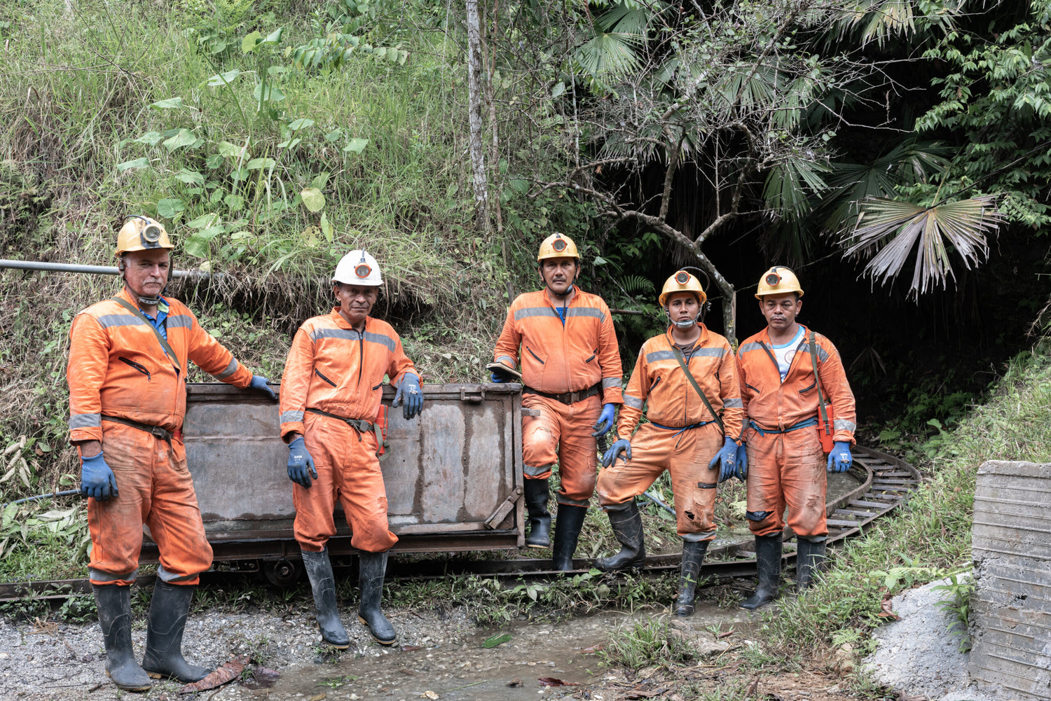 Five workers of fairmined gold in orange safety uniforms and hard hats standing outdoors beside a metal cart on a narrow track, with dense green vegetation in the background.