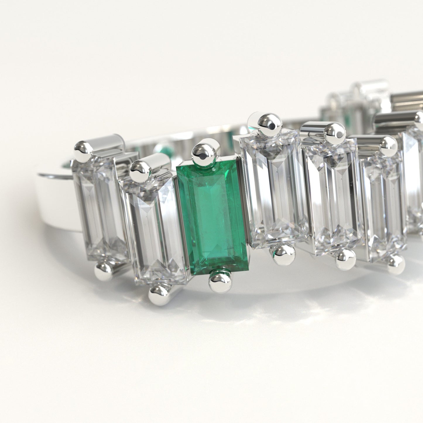 zoom in of a white gold ring featuring a row of clear baguette-cut diamonds with a prominent baguette-cut colombian emerald on the left and a small round green stone near the back, set against a plain white background.