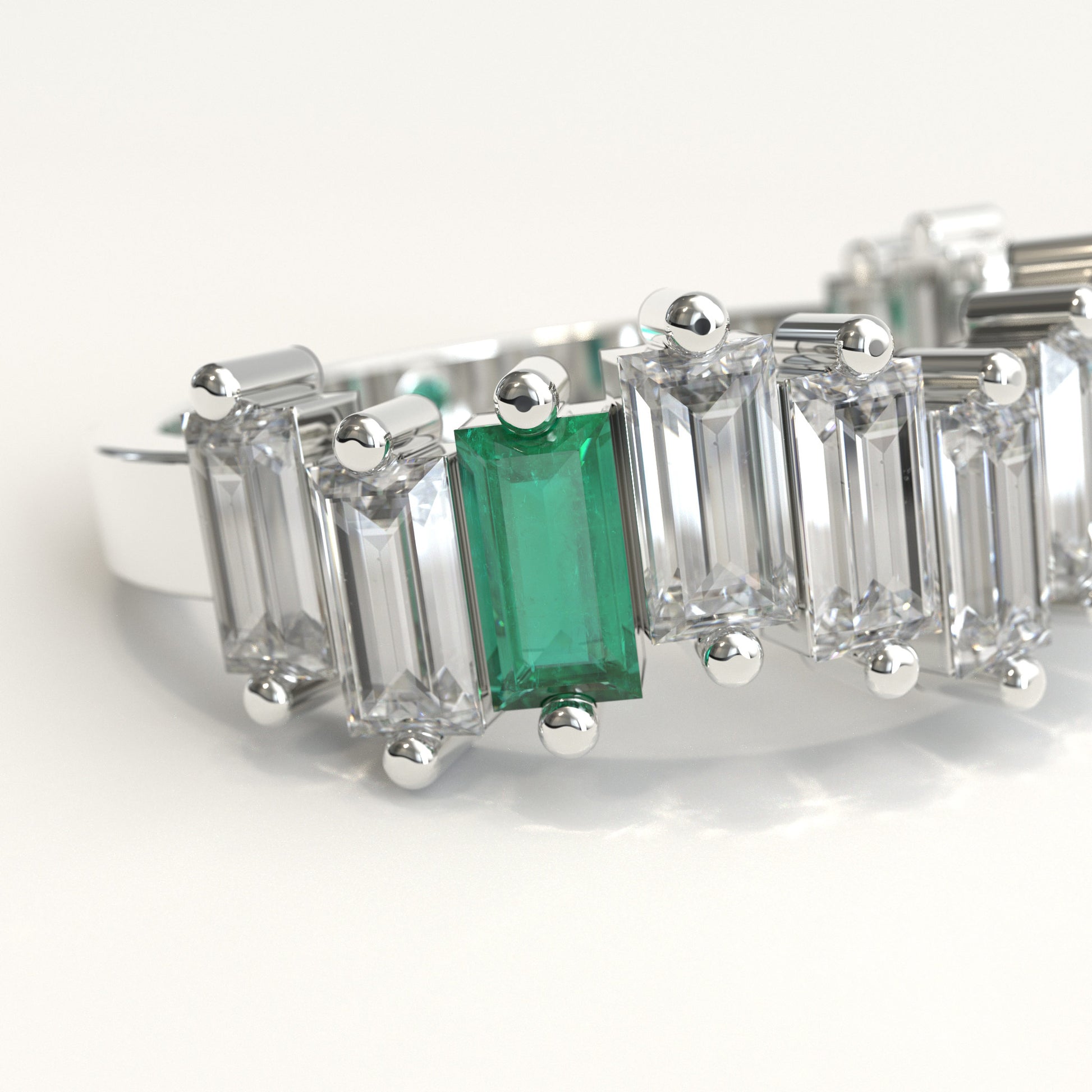 zoom in of a white gold ring featuring a row of clear baguette-cut diamonds with a prominent baguette-cut colombian emerald on the left and a small round green stone near the back, set against a plain white background.