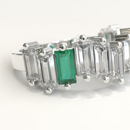 zoom in of a white gold ring featuring a row of clear baguette-cut diamonds with a prominent baguette-cut colombian emerald on the left and a small round green stone near the back, set against a plain white background.