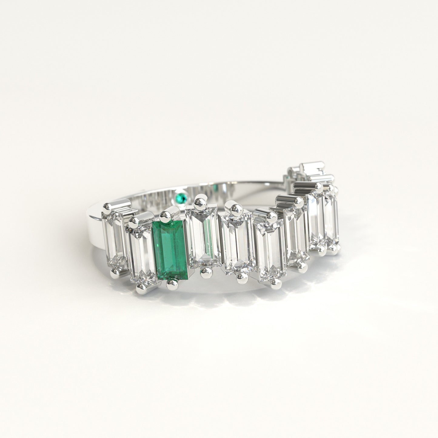 lateral view of a white gold ring featuring a row of clear baguette-cut diamonds with a prominent baguette-cut colombian emerald on the left and a small round green stone near the back, set against a plain white background.