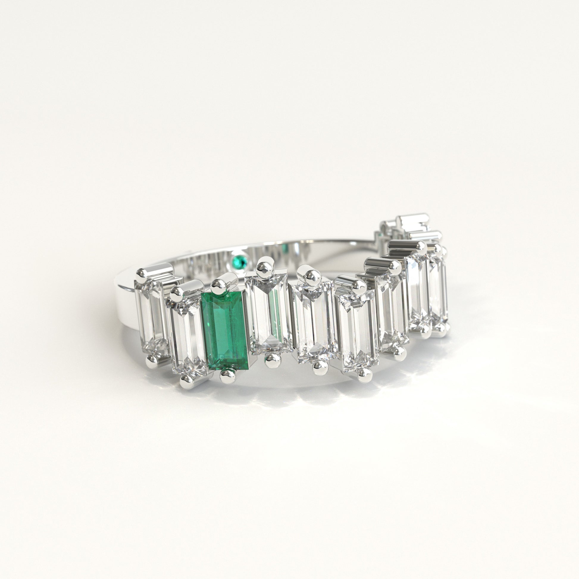 lateral view of a white gold ring featuring a row of clear baguette-cut diamonds with a prominent baguette-cut colombian emerald on the left and a small round green stone near the back, set against a plain white background.