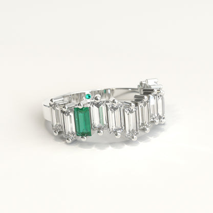 lateral view of a white gold ring featuring a row of clear baguette-cut diamonds with a prominent baguette-cut colombian emerald on the left and a small round green stone near the back, set against a plain white background.