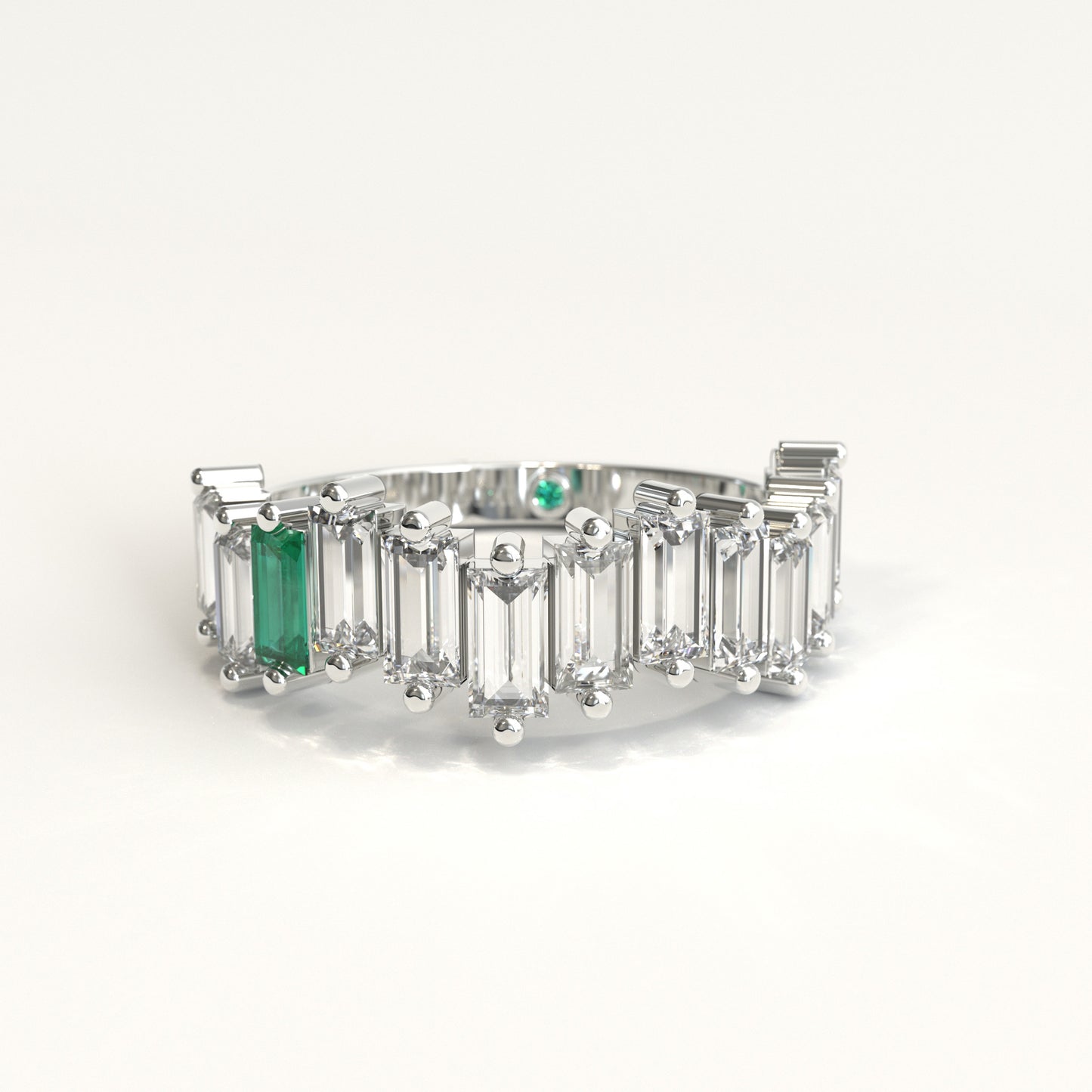 A white gold ring featuring a row of clear baguette-cut diamonds with a prominent baguette-cut colombian emerald on the left and a small round green stone near the back, set against a plain white background.