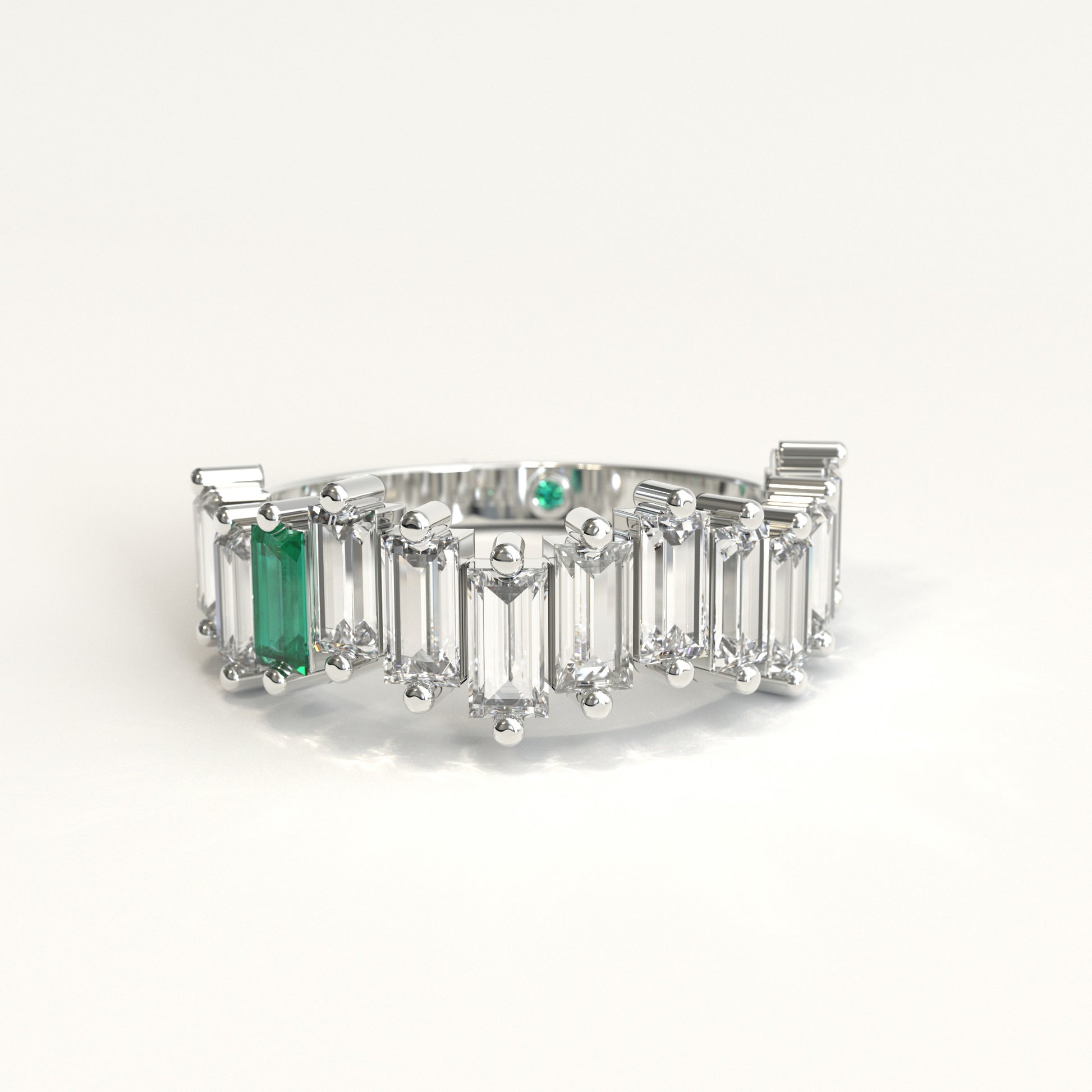 A white gold ring featuring a row of clear baguette-cut diamonds with a prominent baguette-cut colombian emerald on the left and a small round green stone near the back, set against a plain white background.