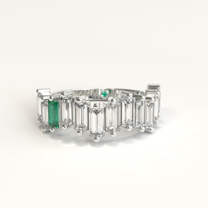 A white gold ring featuring a row of clear baguette-cut diamonds with a prominent baguette-cut colombian emerald on the left and a small round green stone near the back, set against a plain white background.