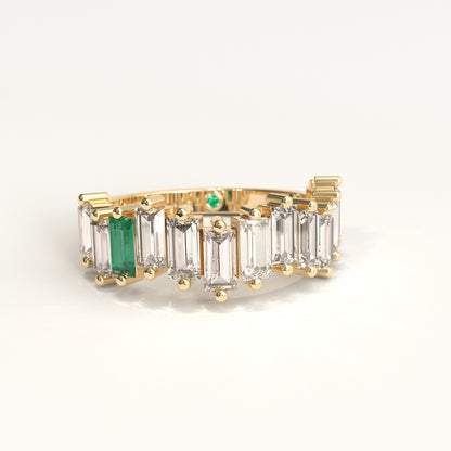 A yellow gold ring featuring a row of baguette-cut ciamonds and one baguette-cut colombian emerald, with a small round green gemstone on the inner band, set against a plain white background.