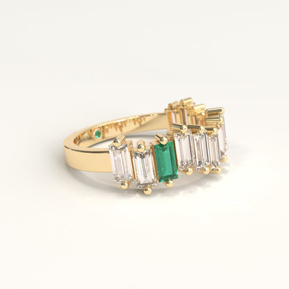 Lateral view of  yellow gold ring featuring a row of baguette-cut ciamonds and one baguette-cut colombian emerald, with a small round green gemstone on the inner band, set against a plain white background.