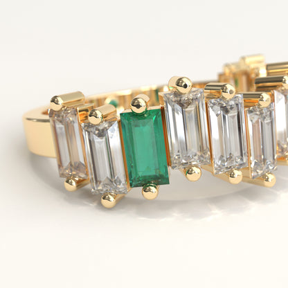 Zoom in of a yellow gold ring featuring a row of baguette-cut ciamonds and one baguette-cut colombian emerald, with a small round green gemstone on the inner band, set against a plain white background.