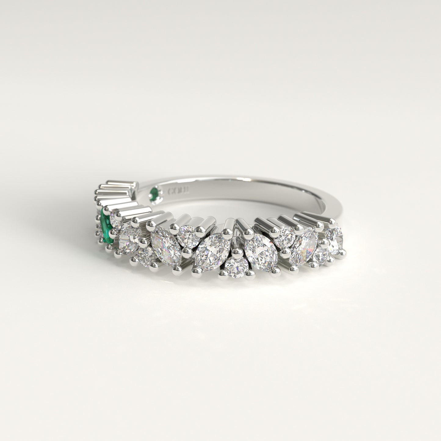A white 18k gold band ring featuring a row of diamonds, including marquise-cut, and round-cut stones, accented by two small green emerald-cut stones on the left side, set against a clean white background.