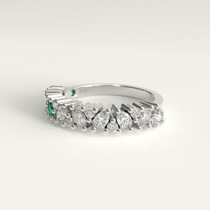 A white 18k gold band ring featuring a row of diamonds, including marquise-cut, and round-cut stones, accented by two small green emerald-cut stones on the left side, set against a clean white background.