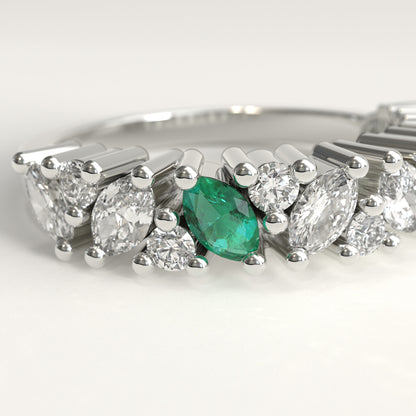 Datail of a 18k  white gold band ring featuring a row of diamonds, including marquise-cut, and round-cut stones, accented by two small green emerald-cut stones on the left side, set against a clean white background.