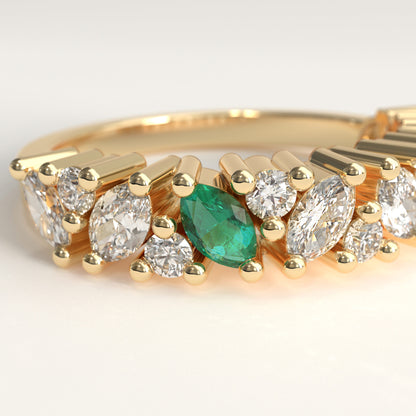 close up of a yellow gold eternity-style ring featuring a row of round and marquise-cut diamonds with a central green marquise cut emerald, photographed on a clean light gray background.