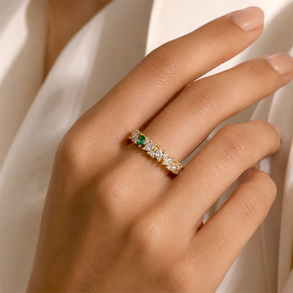 A close-up of a hand wearing a delicate gold eternity ring with a single colombian emerald and diamonds, set against a soft white draped fabric background.