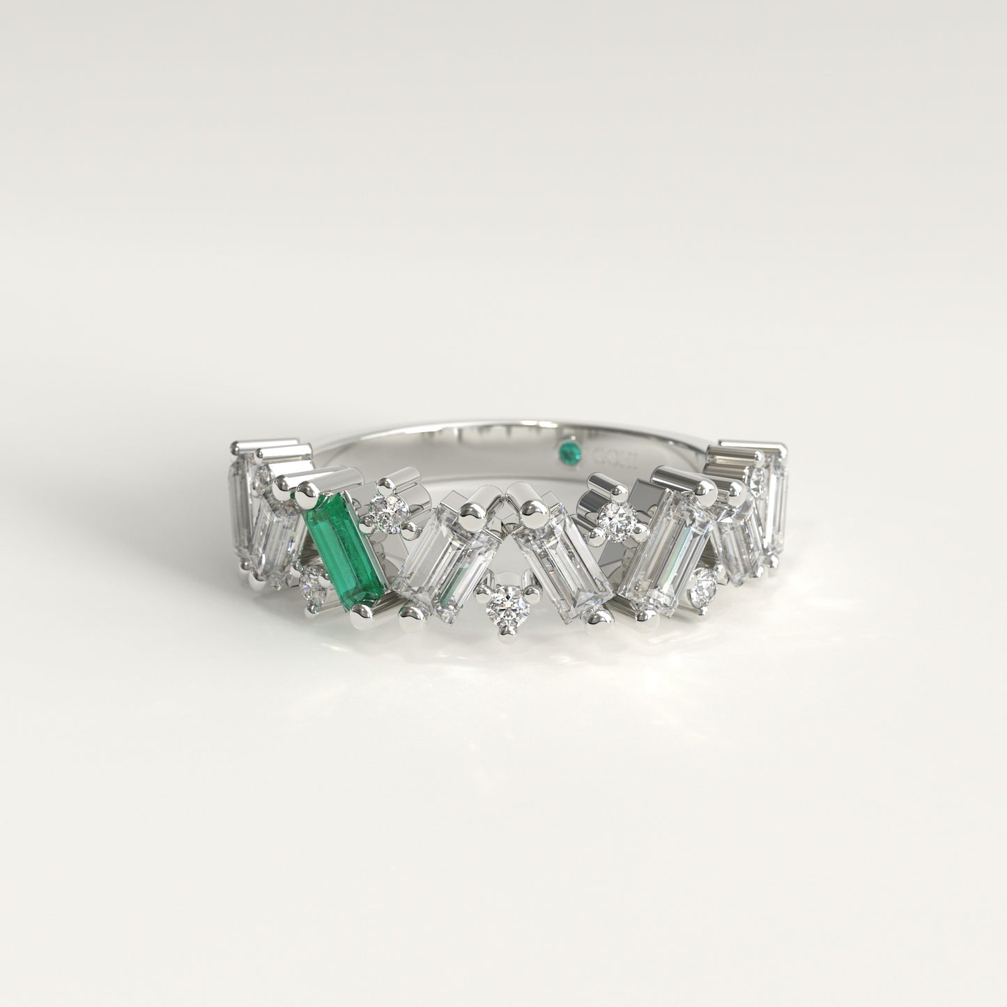 A white 18k fairmined gold diamond band ring featuring a row of baguette and round-cut clear diamonds with a prominent emerald baguette-cut gemstone on the left side, set against a clean white background.