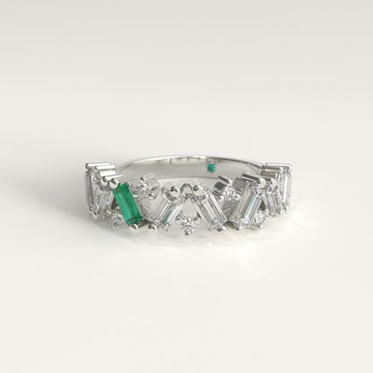 A white 18k fairmined gold diamond band ring featuring a row of baguette and round-cut clear diamonds with a prominent emerald baguette-cut gemstone on the left side, set against a clean white background.