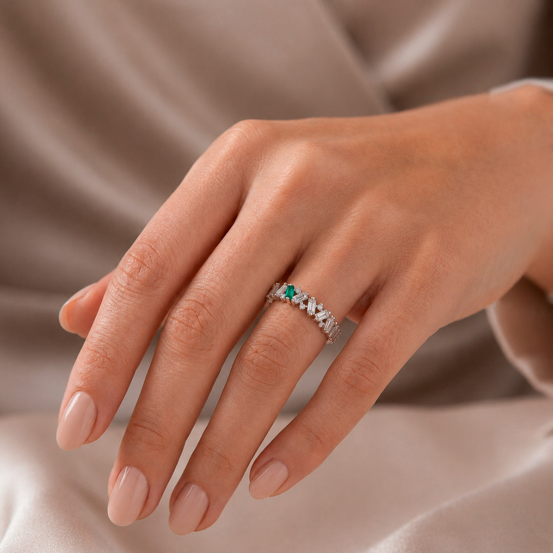 A close-up of a hand wearing a white gold ring with a single Colombian Emerald baguette-cut center stone and small Diamonds along the band, set against a soft beige satin background.