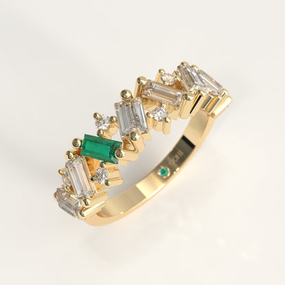 A gold band ring featuring a row of baguette and round diamonds, accented by a prominent colombian emerald baguette-cut and a small round green stone on the band, shown on a light gray background.
