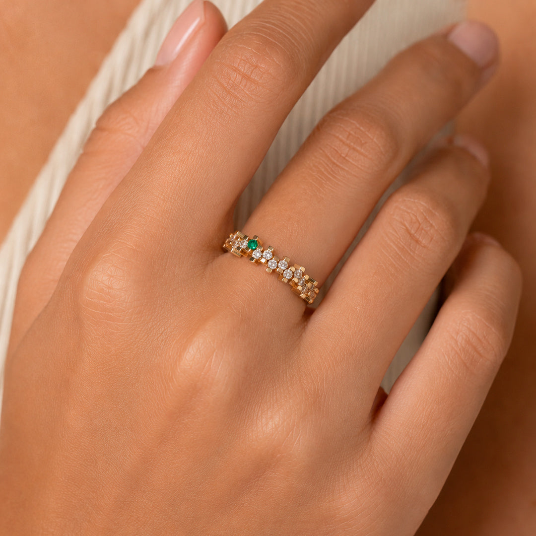A close-up of a hand wearing a delicate yellow gold ring with a geometric design, set with round white diamonds and one vibrant Colombian emerald. The warm gold, brilliant diamonds, and vivid green gemstone create a refined, modern look.