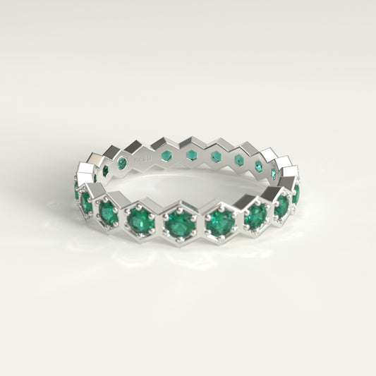 A 18k fairmined white gold eternity ring featuring a repeating geometric hexagonal pattern set with round green emeralds, shown on a clean light gray background.
