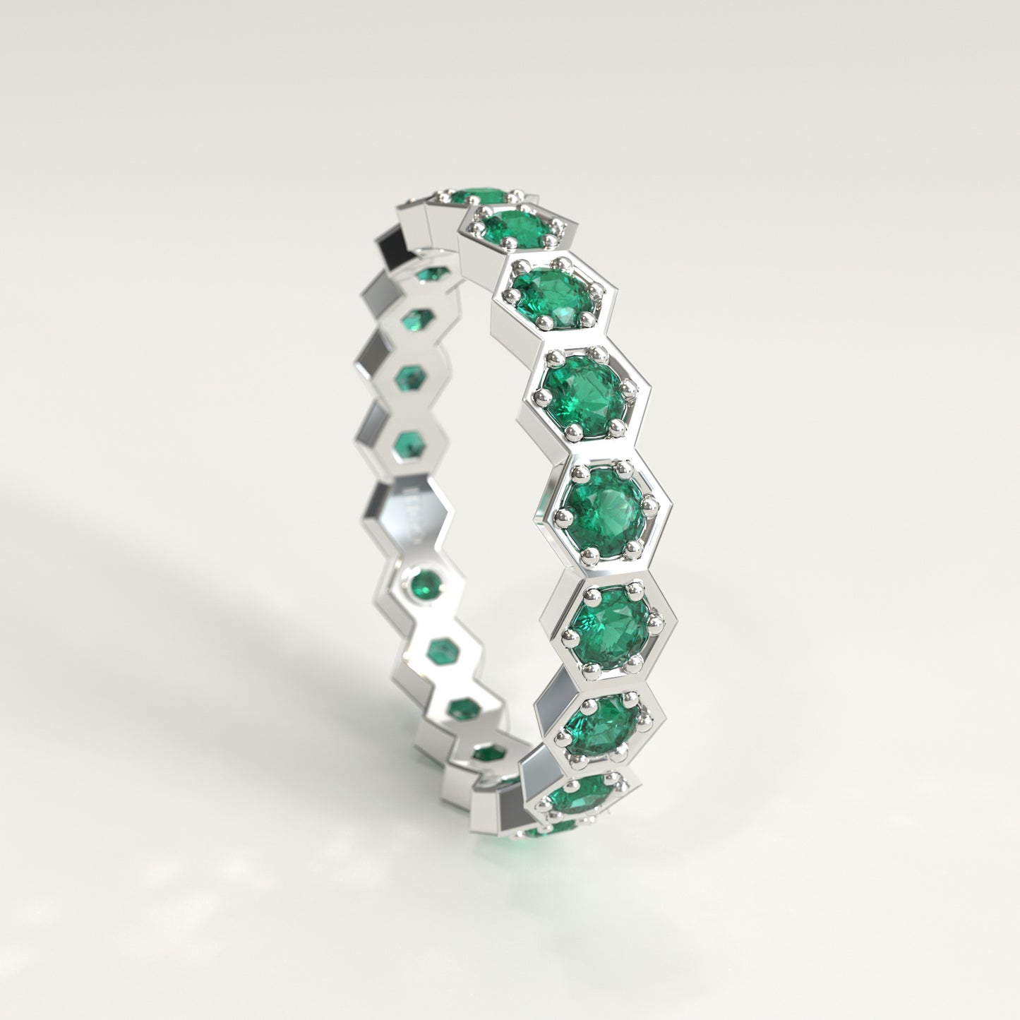 A white gold honeycomb eternity ring set with round green emeralds, shown on a clean light gray background.