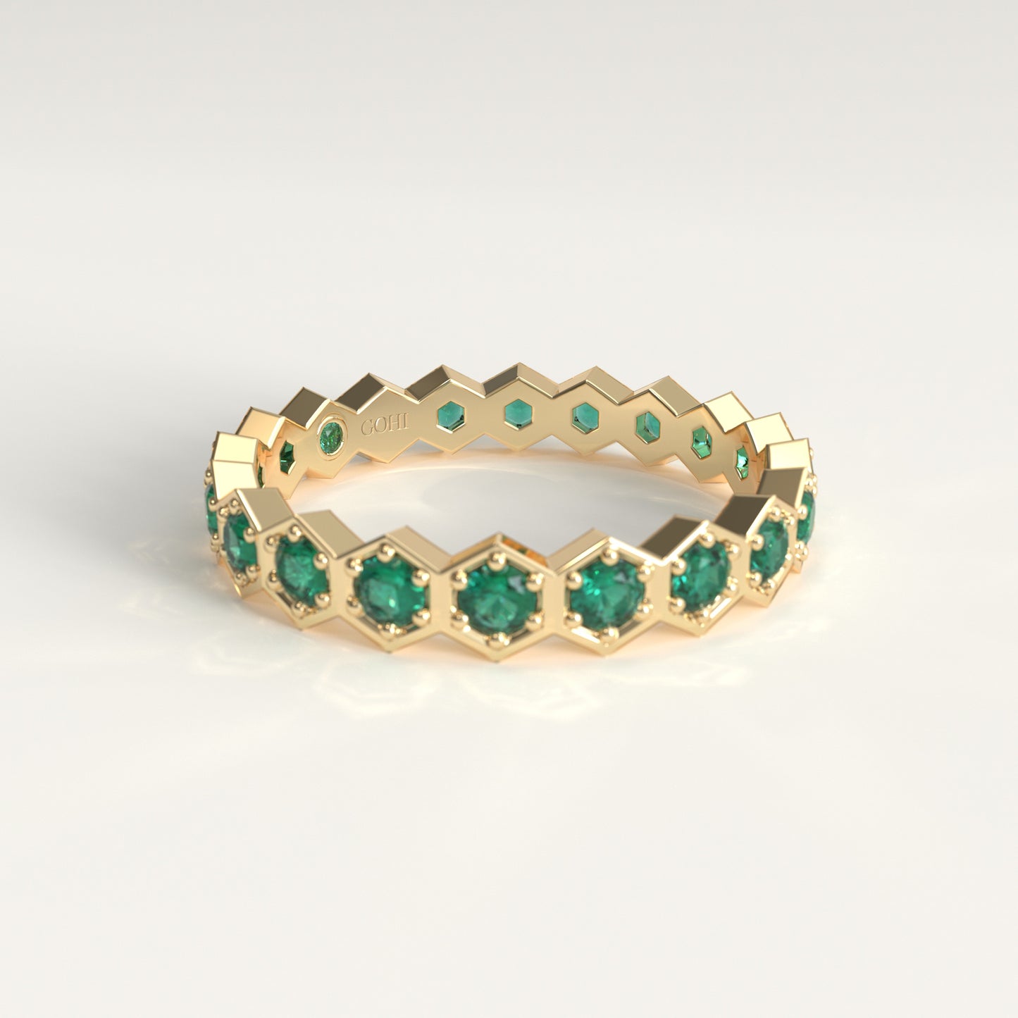 A gold eternity ring featuring a repeating hexagonal pattern with round-cut green emerald gemstones, shown on a clean white background.