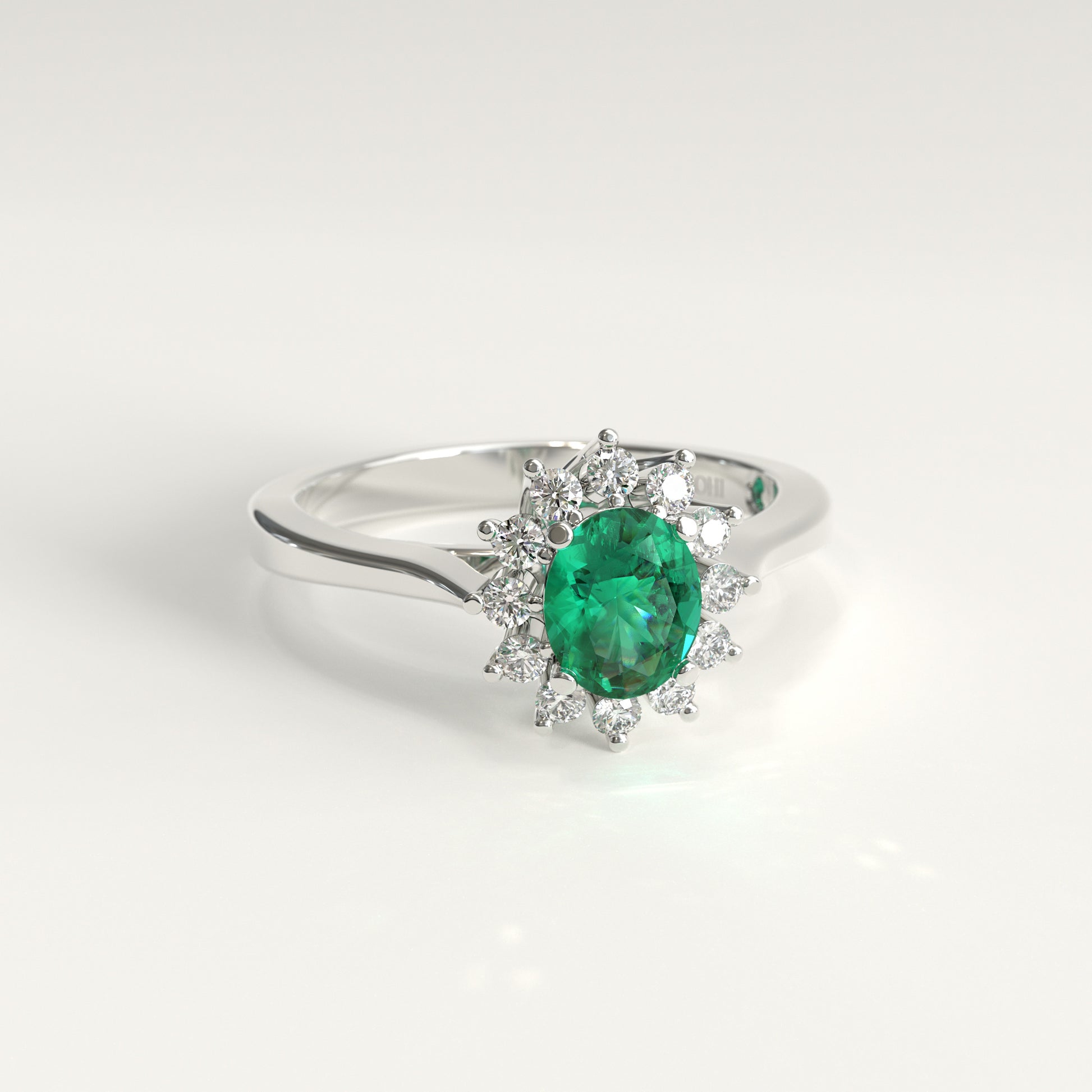 An engagement ring in 18k white gold. A green oval-cut Colombian emerald sits at the center, surrounded by twelve small round diamonds arranged in a flower-like halo. The band tapers gently upward on each side to meet the setting. GOHI Bloom Ring, Engagement Collection.