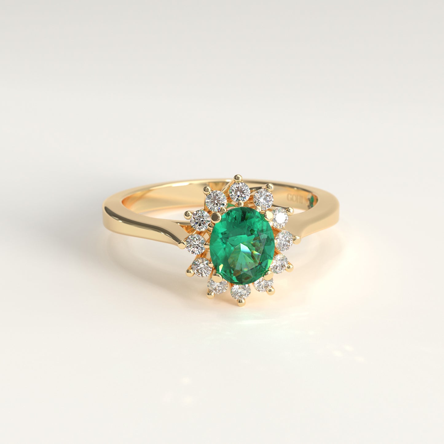 An engagement ring in 18k yellow gold. A green oval-cut Colombian emerald sits at the center, surrounded by twelve small round diamonds arranged in a flower-like halo. The band tapers gently upward on each side to meet the setting. GOHI Bloom Ring, Engagement Collection.