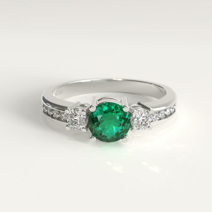 An engagement ring in 18k white gold. A round green Colombian emerald sits at the center, flanked by one round white diamond on each side, each held by its own set of prongs. Smaller diamonds line the band on both shoulders. GOHI Trilogy Ring, Engagement Collection.