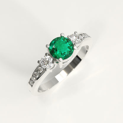 A 18k fairmined gold ring featuring a round-cut green colombian emerald center stone and small diamonds accent stones on the shoulders, set against a plain white background.