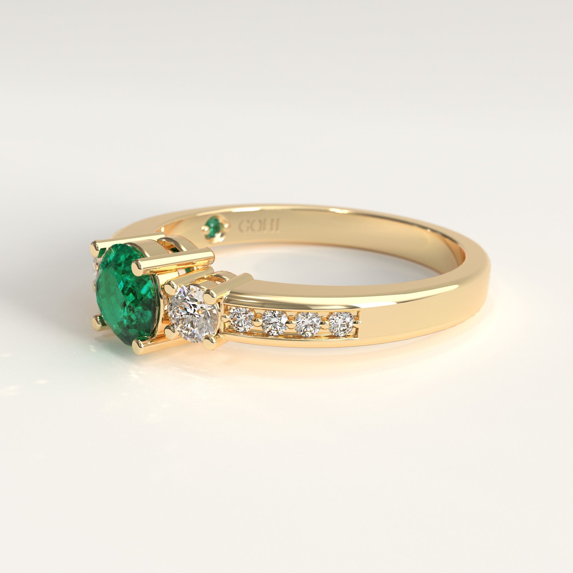 lateral view of a Trilogy Ring crafted in 18k yellow gold — a round green Colombian emerald at the center, flanked by two round diamonds, with smaller diamonds set along both shoulders.