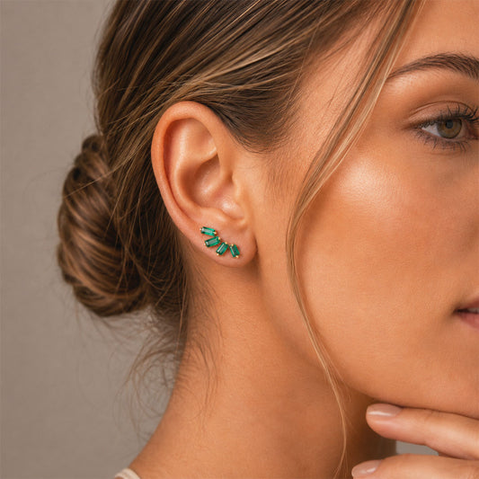 A close-up of a woman's ear wearing a small cluster stud earring with multiple baguette-cut colombian emeralds set in yellow gold, against a soft neutral background.
