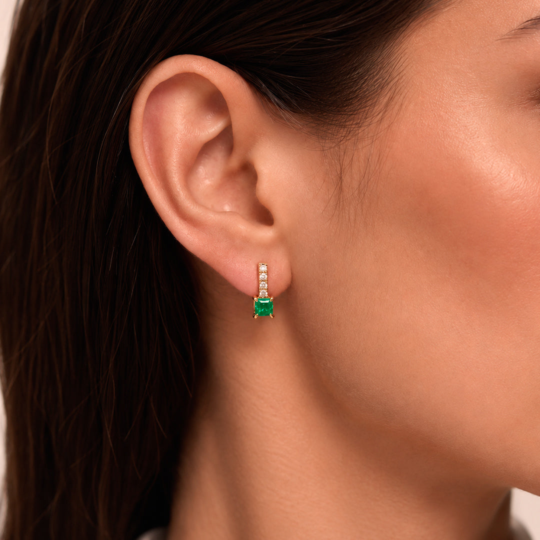 A close-up of a model's ear wearing a small drop earring with a rectangular Colombian Emerald and a row of 4 diamonds above it.