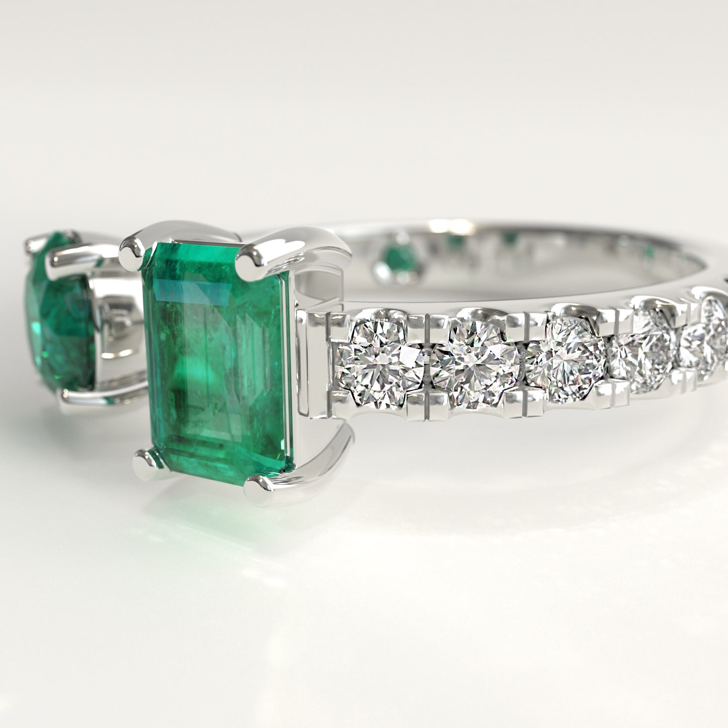 Close-up of round and emerald-cut Colombian emeralds on open toi et moi ring, Duet Ring by GOHI Soiree Collection. A white gold diamond ring featuring a large emerald-cut green emerald and a smaller matching green stone on the shoulders, with a row of clear round diamonds along the band, set against a clean white background.