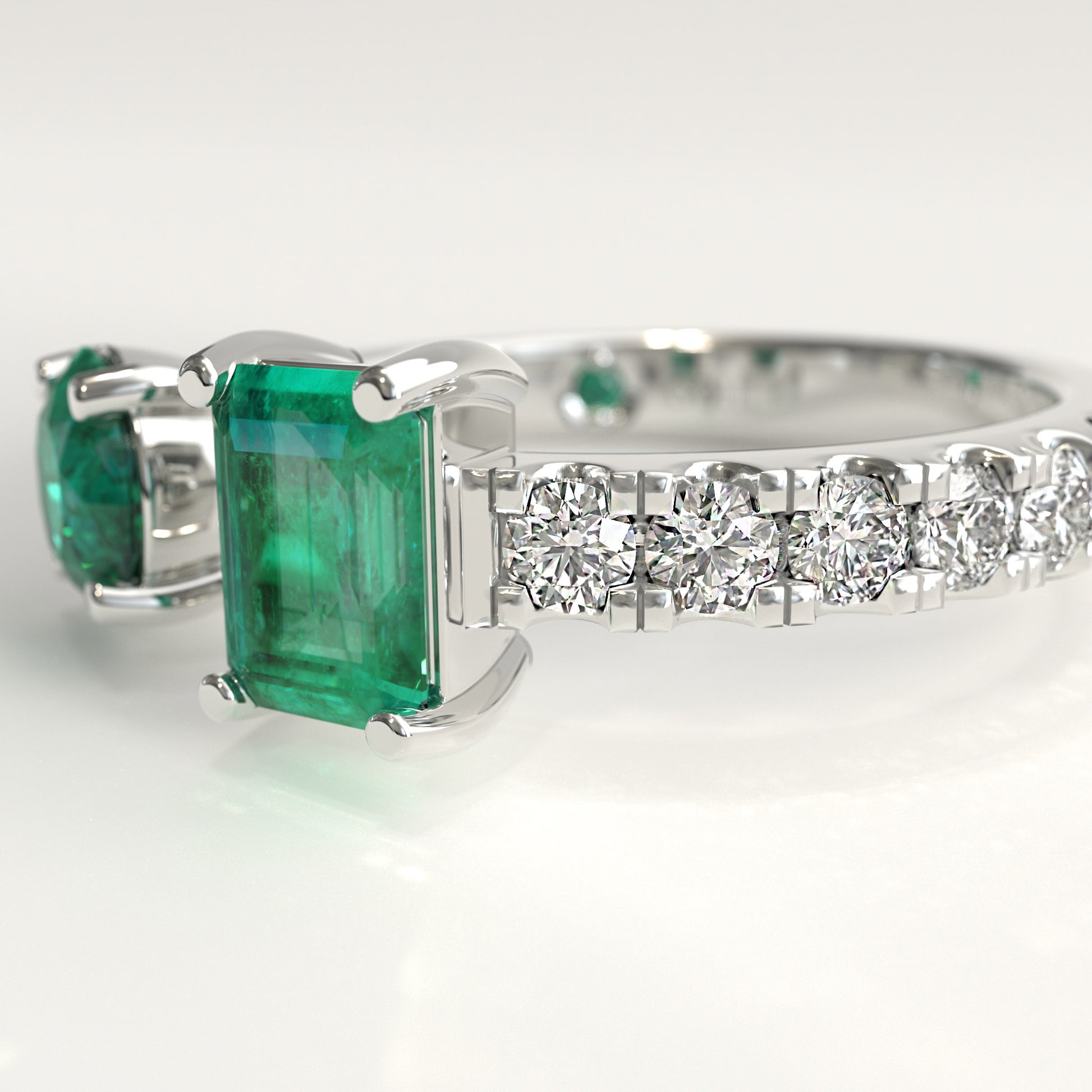 Close-up of round and emerald-cut Colombian emeralds on open toi et moi ring, Duet Ring by GOHI Soiree Collection. A white gold diamond ring featuring a large emerald-cut green emerald and a smaller matching green stone on the shoulders, with a row of clear round diamonds along the band, set against a clean white background.