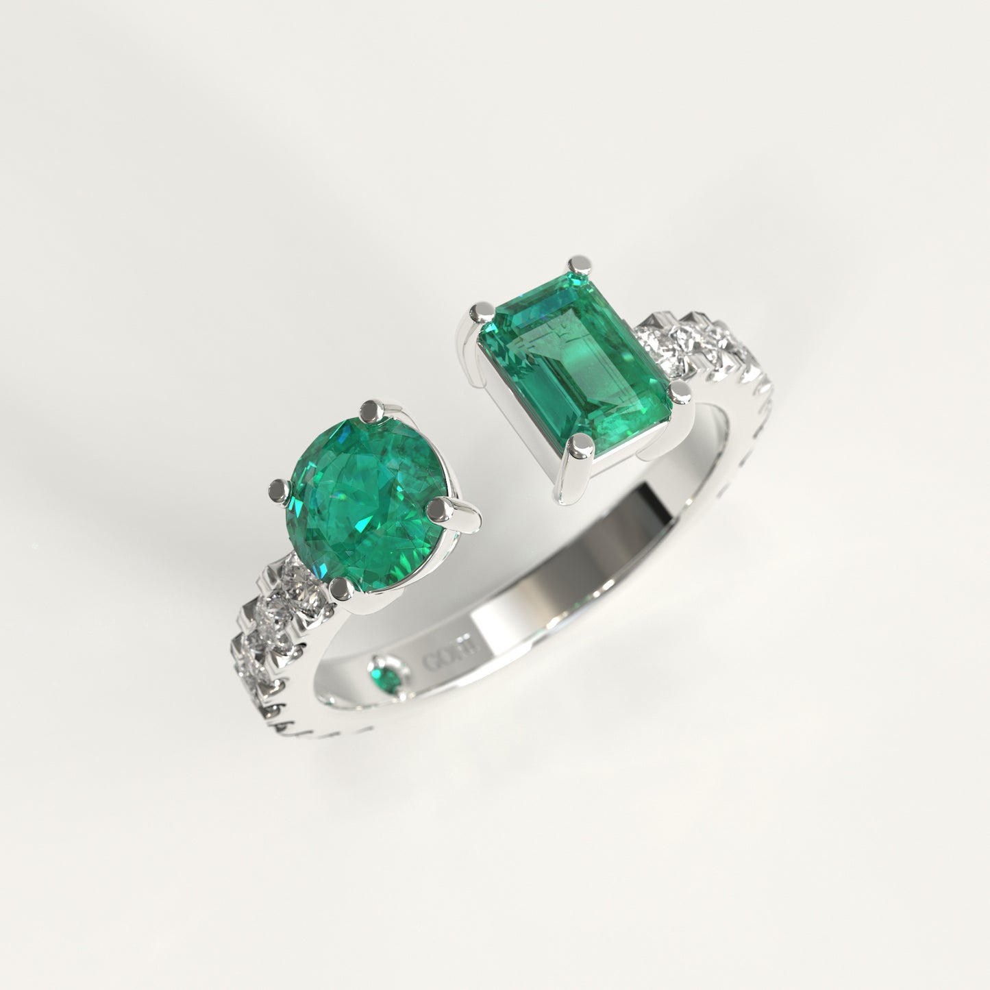 A white gold ring featuring a large round-cut colombian green emerald and a rectangular emerald-cut colombian emerald, accented with small diamonds on the band, shown on a light gray background.