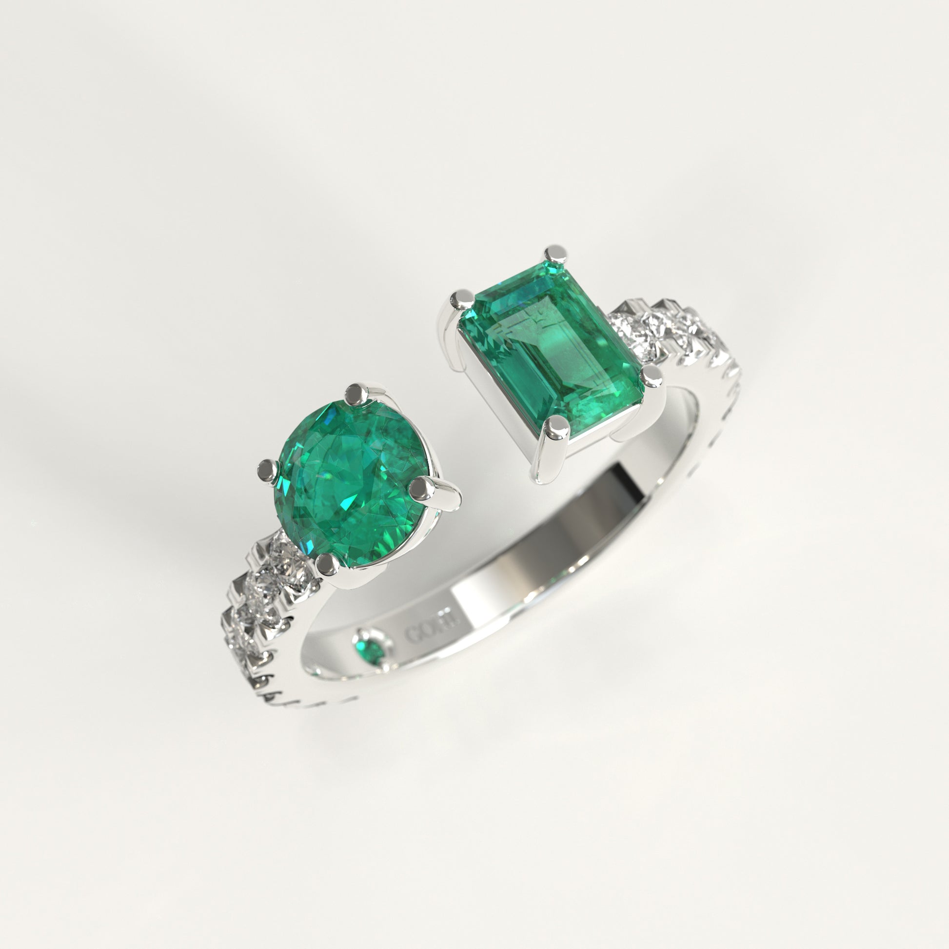 A white gold ring featuring a large round-cut colombian green emerald and a rectangular emerald-cut colombian emerald, accented with small diamonds on the band, shown on a light gray background.