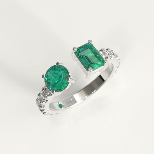 A white gold ring featuring a large round-cut colombian green emerald and a rectangular emerald-cut colombian emerald, accented with small diamonds on the band, shown on a light gray background.