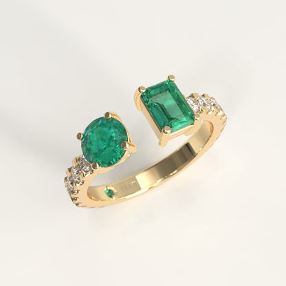 A 18k fairmined gold ring featuring a large round-cut colombian green emerald and an emerald-cut colombain emerald, accented with small diamonds on the shoulders, shown on a light gray background.