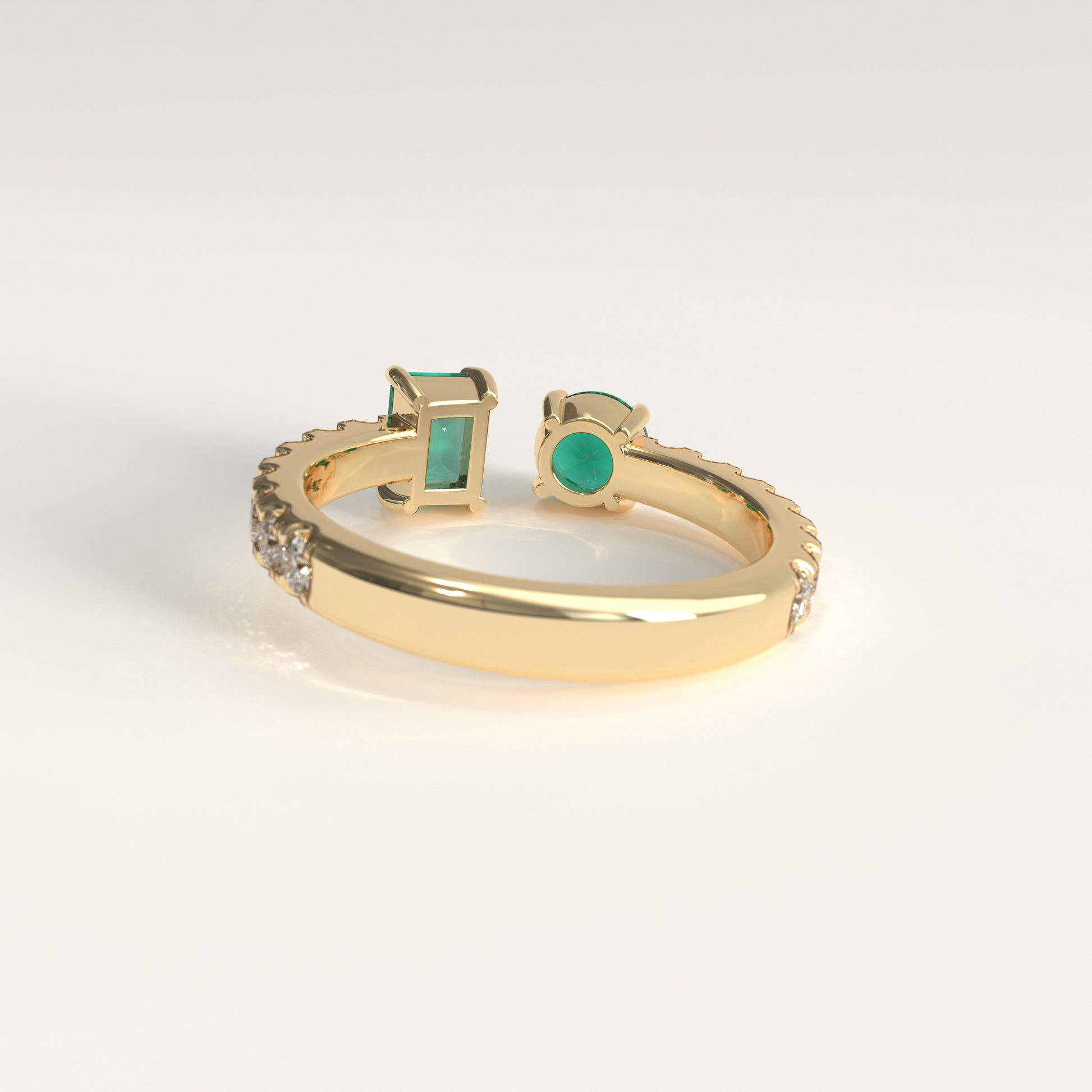 Back view of Duet Ring showing hidden Colombian emerald beside GOHI signature, 18k yellow gold. A gold open-shank ring with a rectangular emerald-cut colombian emerald on one side and a round green emerald on the other, accented with small diamonds along the band, shown on a light gray background.