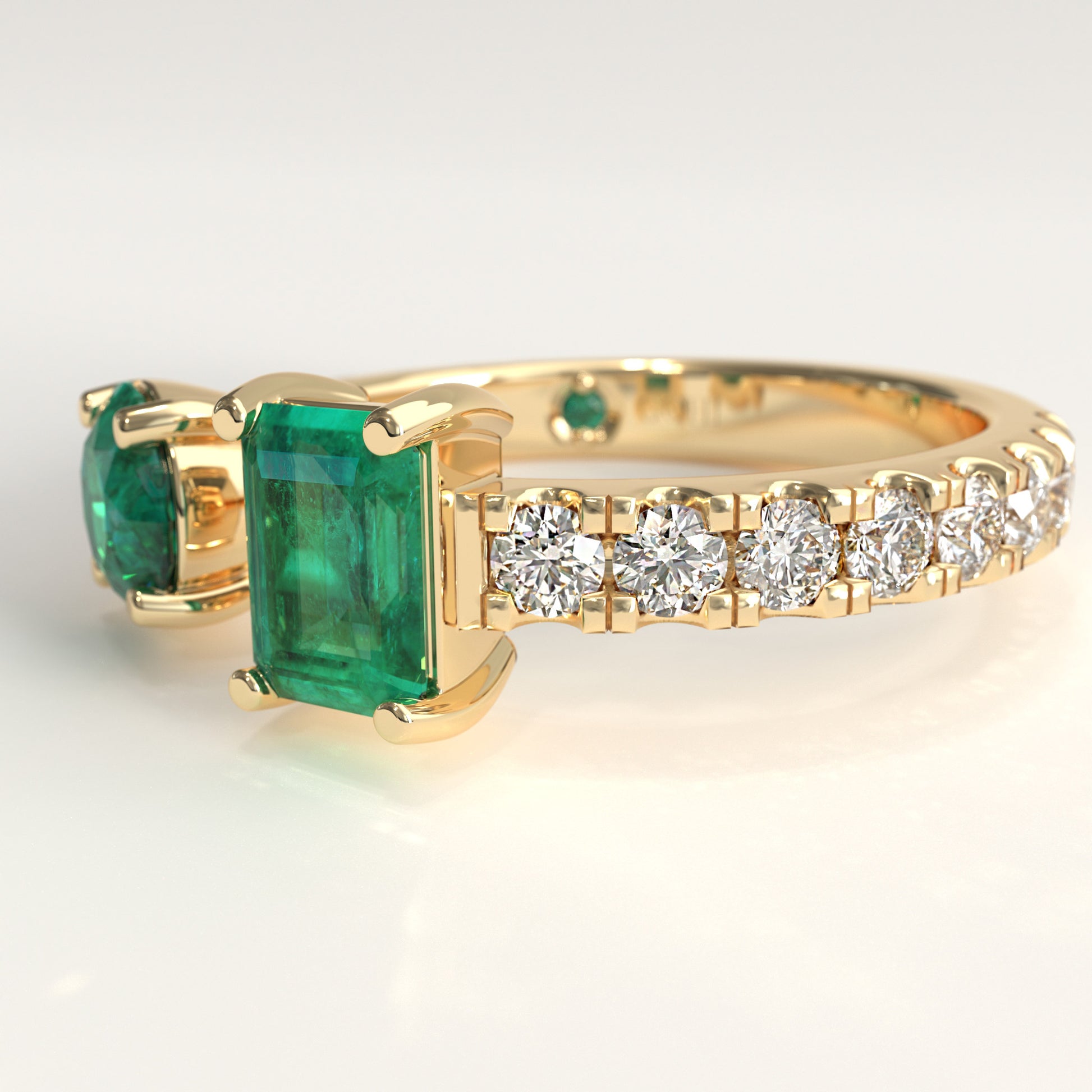 Close-up of round and emerald-cut Colombian emeralds on open toi et moi ring, Duet Ring by GOHI Soiree Collection. A 18k fairmiend yellow gold ring featuring a large rectangular emerald-cut Colombian emerald gemstone, a smaller round emerald-cut colombian emerald on the opposite side, and a row of round brilliant-cut diamonds along the band.