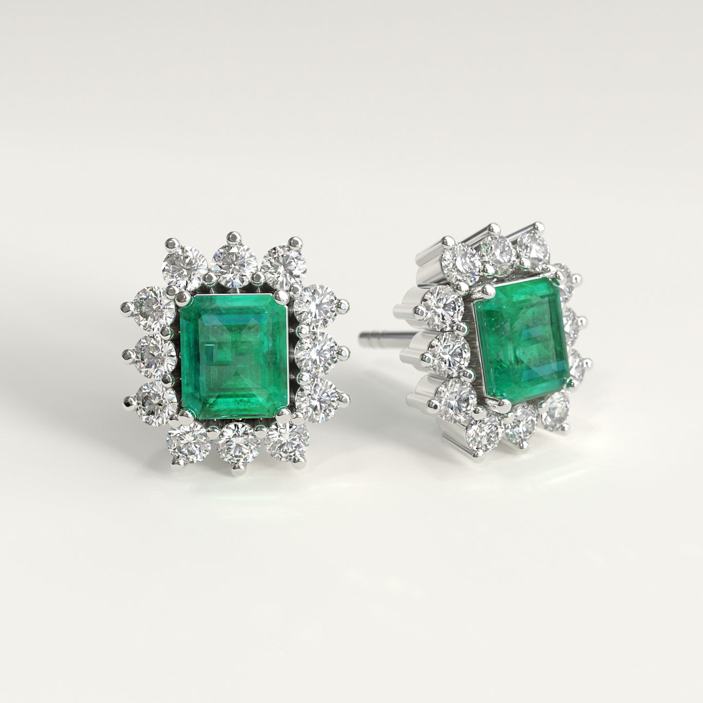 A pair of white gold stud earrings featuring rectangular emerald-cut colombian emerald surrounded by halo settings of small diamonds, shown on a clean white background.